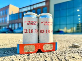 two pale cans sit behind a pair of solar eclipse glasses, outside, with a large set of buildings in the backgound.
