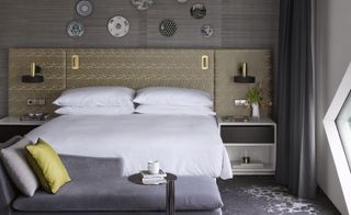 Double bedroom in grey tones, with a bronze headboard with geometric detailing, grey chaise longue and plates hanging on the wall