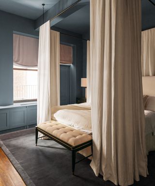 how to make a bedroom darker, blue and cream bedroom with four poster bed with cream linen drapes, blinds, textured cream bedding, grey rug, bench seat, blue ceiling