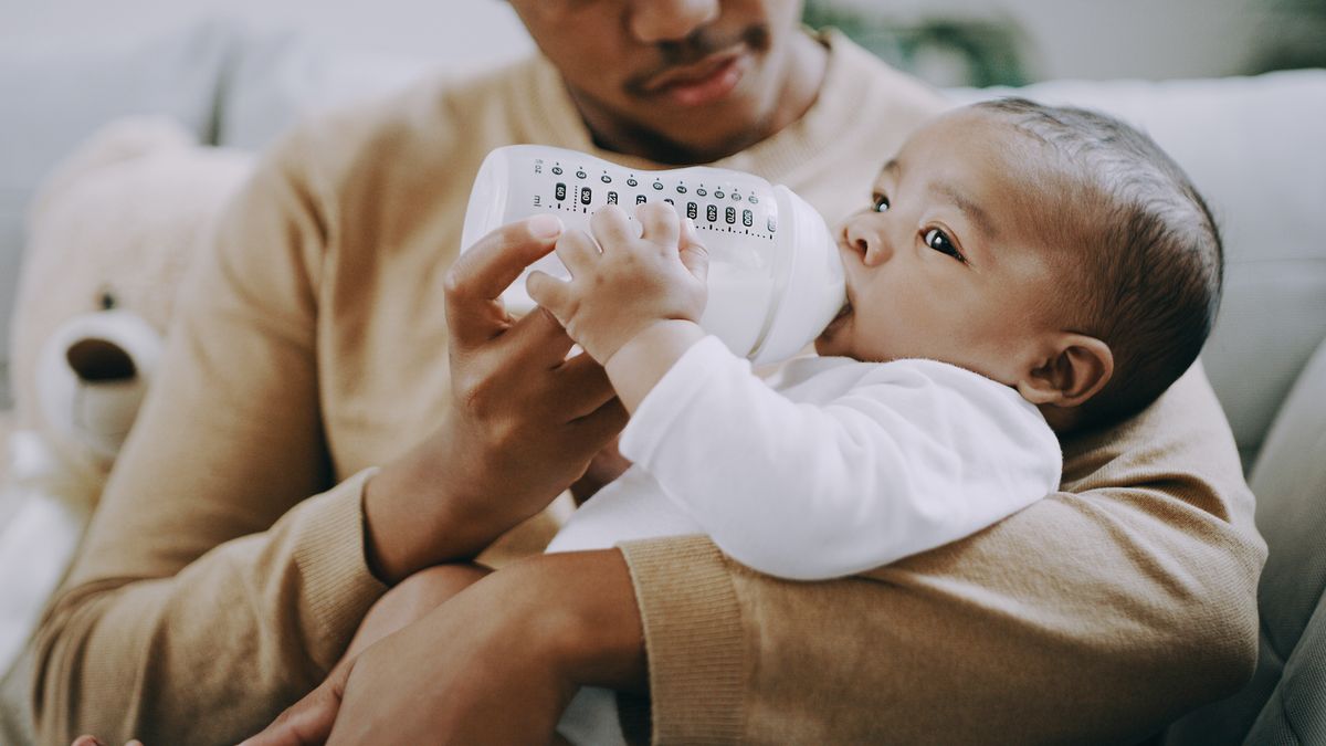 A father sitting on a couch feeds his baby a bottle filled with milk or formula.