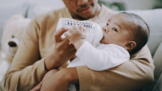 A father sitting on a couch feeds his baby a bottle filled with milk or formula.