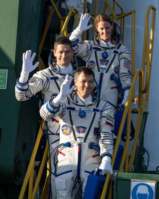 three astronauts in white flight suits wave while standing on a set of stairs.