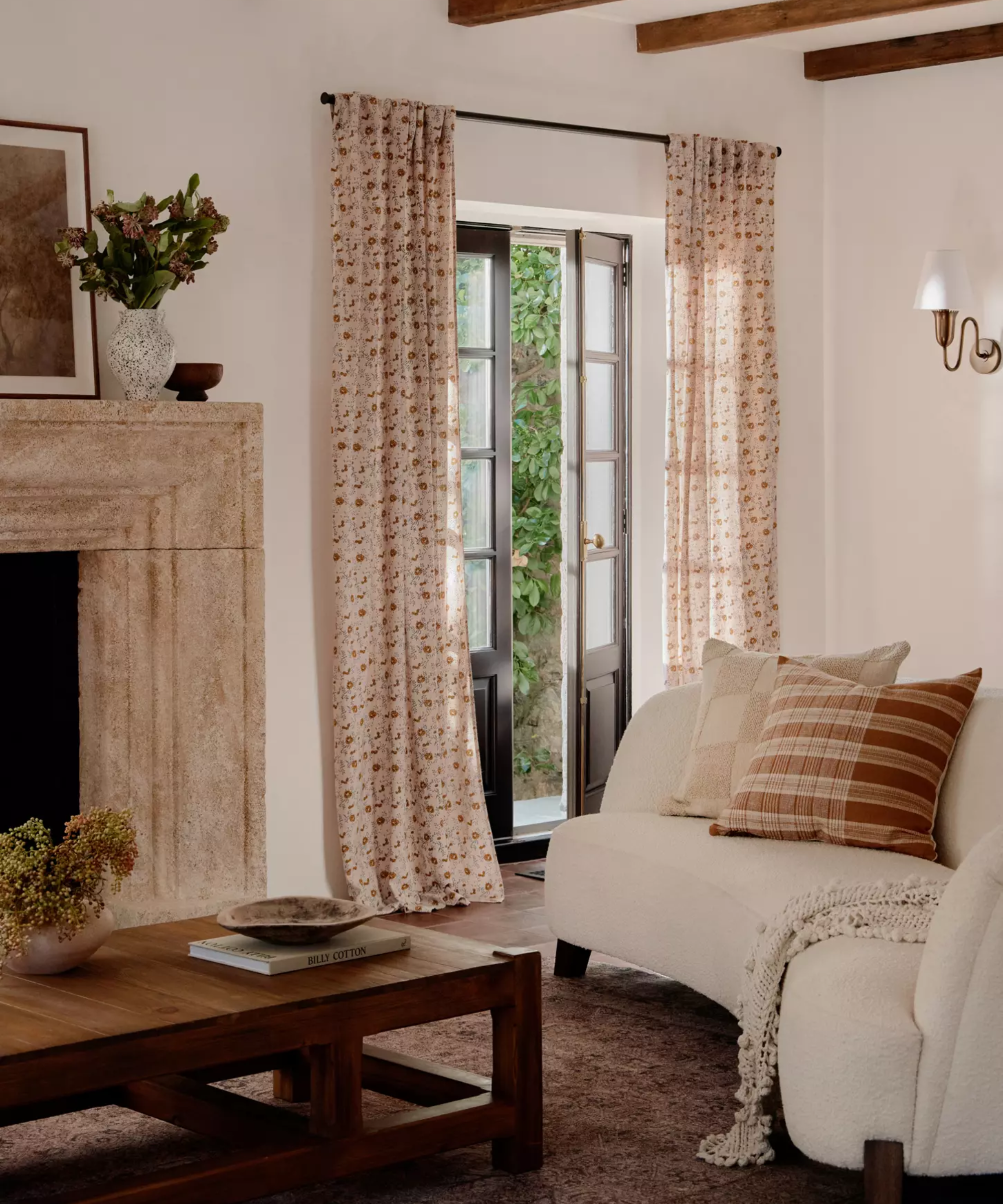a living room with long floral curtains over french doors, a boucle couch, pillows, a wooden coffee table, and a fireplace