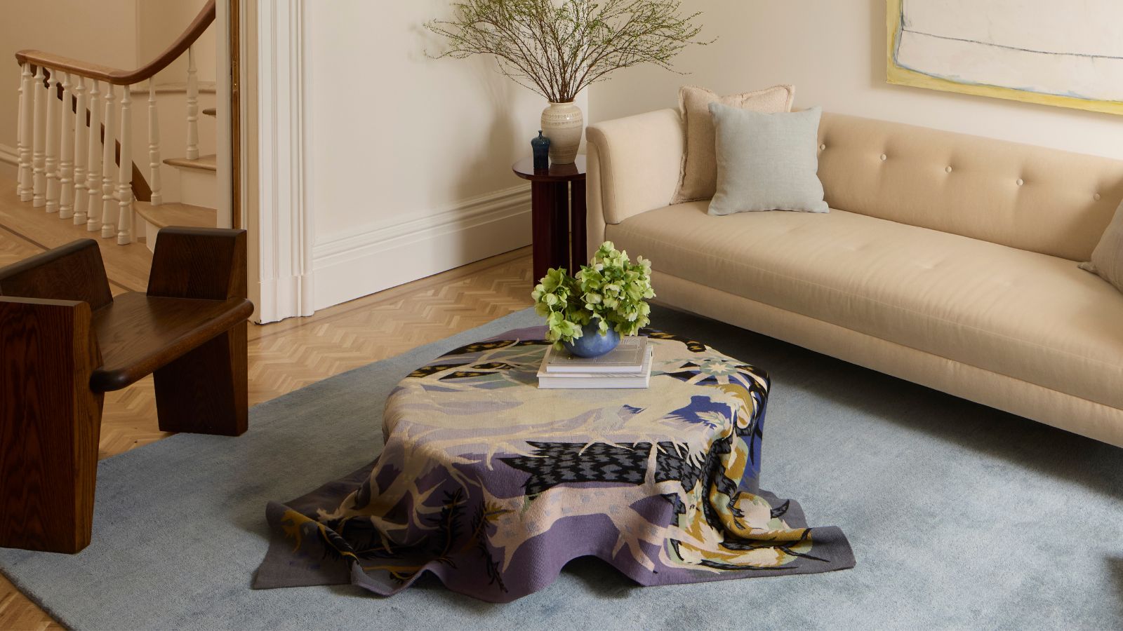 Living room with large beige sofa, coffee table covered in colorful cloth, and a brown wooden chair on a blue rug and parquet wooden floors.