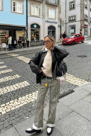 A woman standing near a crosswalk wearing a faded leather jacket over a cream crewneck sweater with a white button-down layered underneath, cheetah pants, and black and white loafers.