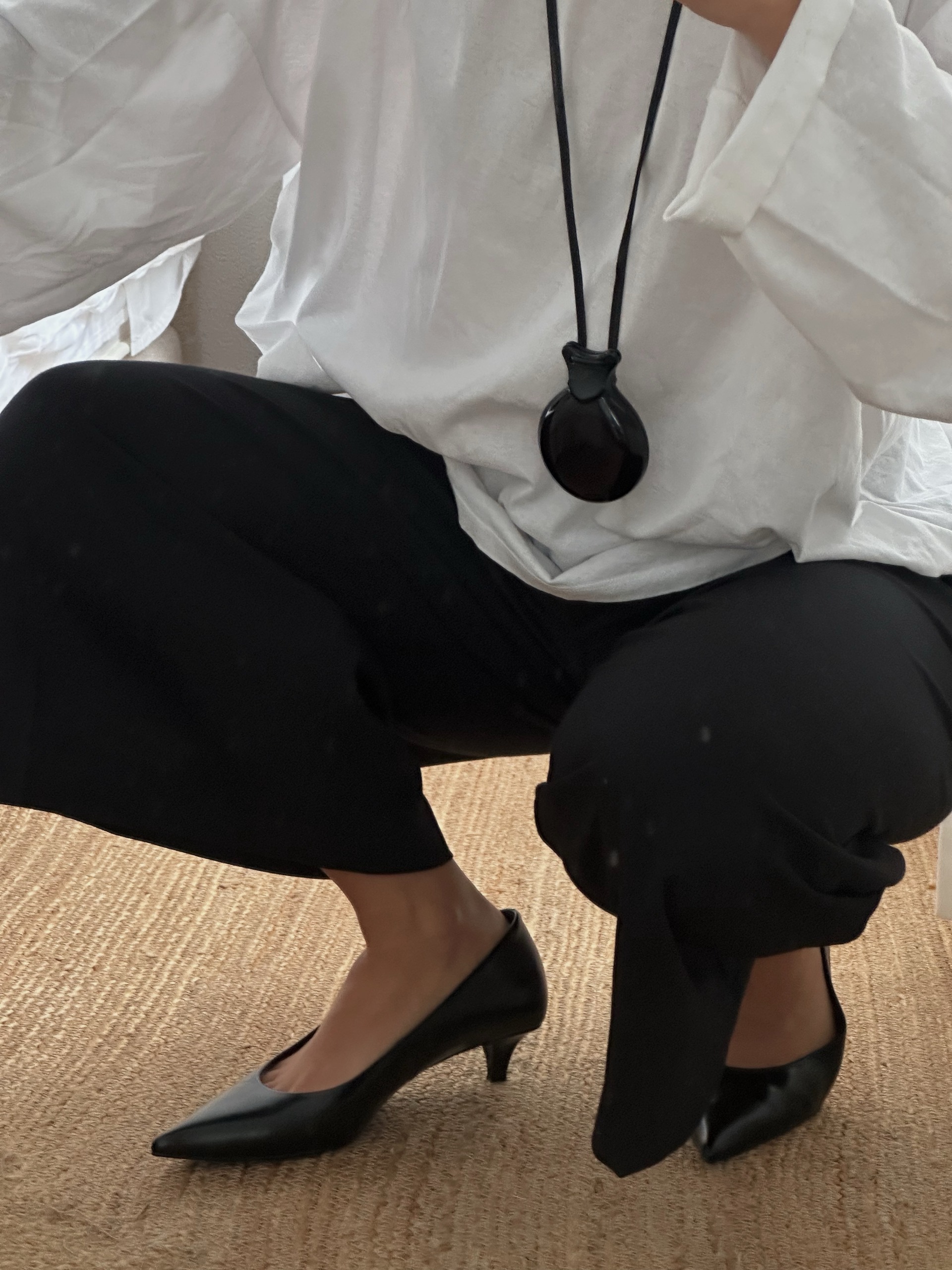 A close up image of an influencer wearing cone heels with black trousers, a white shirt and a black pendant necklace