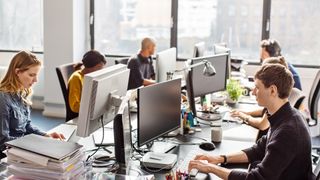 Hybrid work concept image showing a mixture of Gen Z and millennial workers sitting at desks working on computers in an open plan office space.