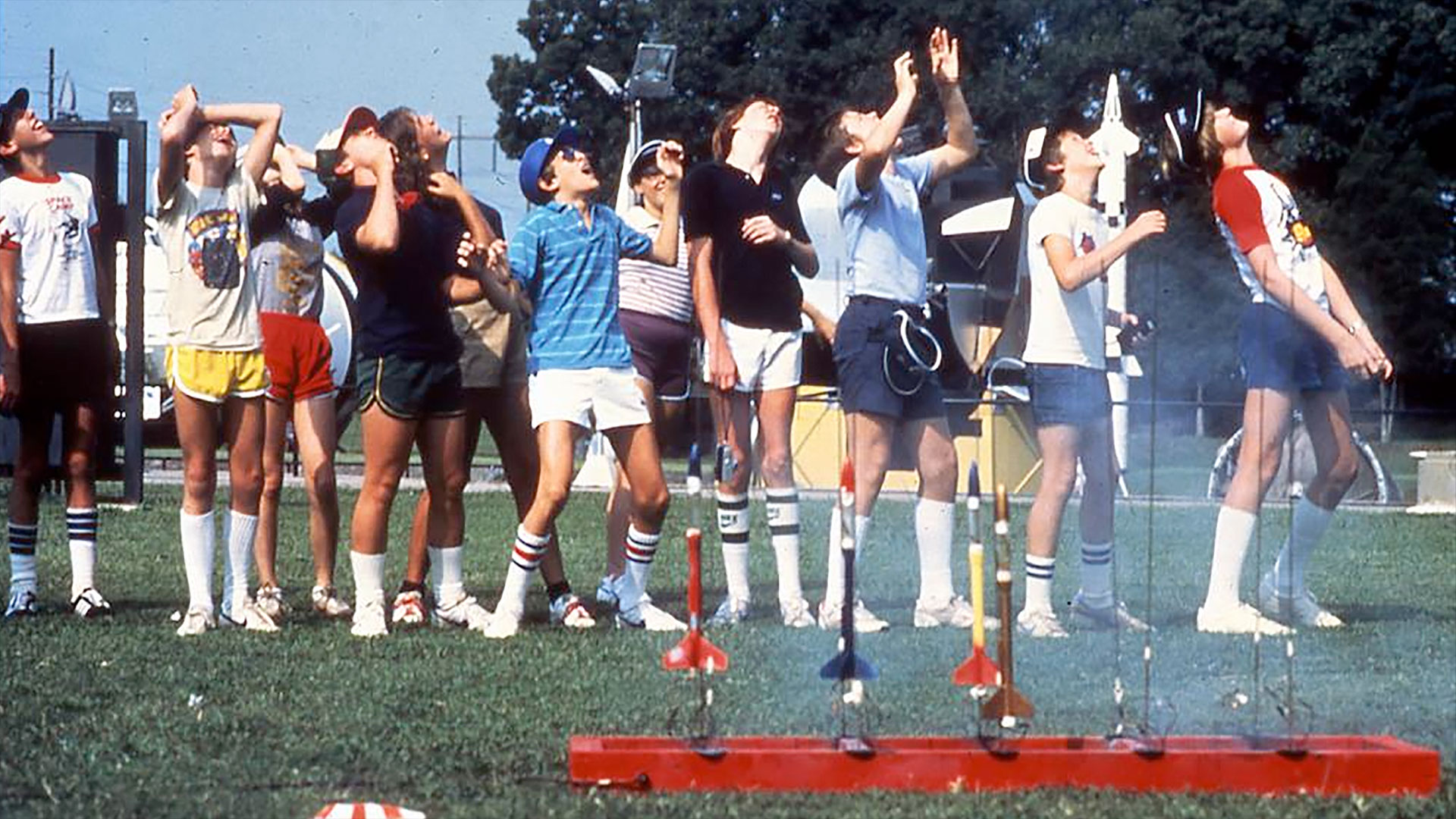 kids in multi-color t-shirts and shorts watch model rockets fly as other rockets stand ready to fly from a launch pad