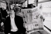 Black and white photograph of passengers on a bus. A gentleman wearing a hat reads a large newspaper whilst the man sat the left tips his head backwards.