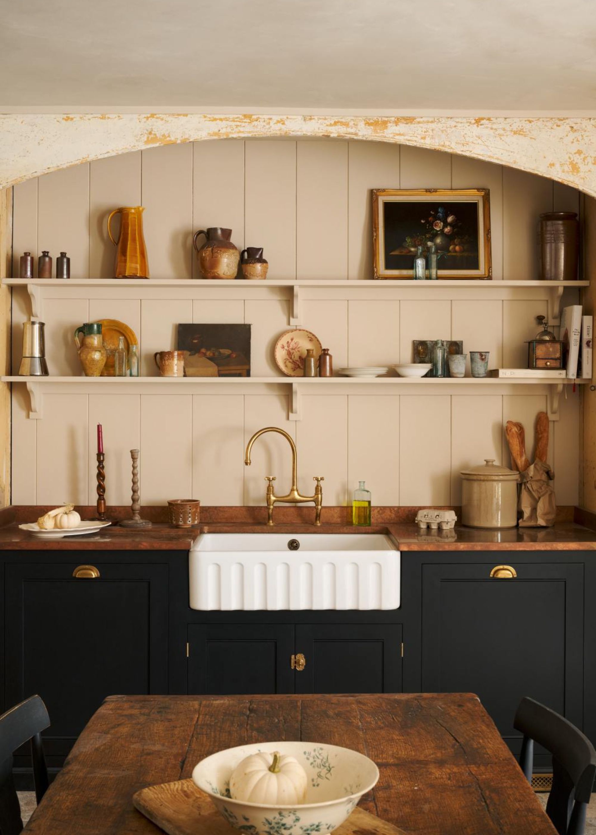 Kitchen cabinets painted black with a wooden countertop and a belfast sink in the middle. Above the sink is open shelving with bowls and artwork