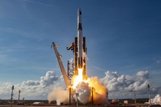 rocket lifts off with clouds in blue sky in behind and orange flame and smoke below