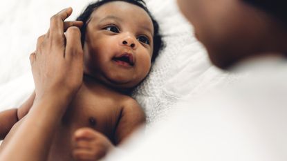 A newborn baby lies on a white blanket, looking up at an adult standing over him. The adult is brushing a strand of the baby's hair back