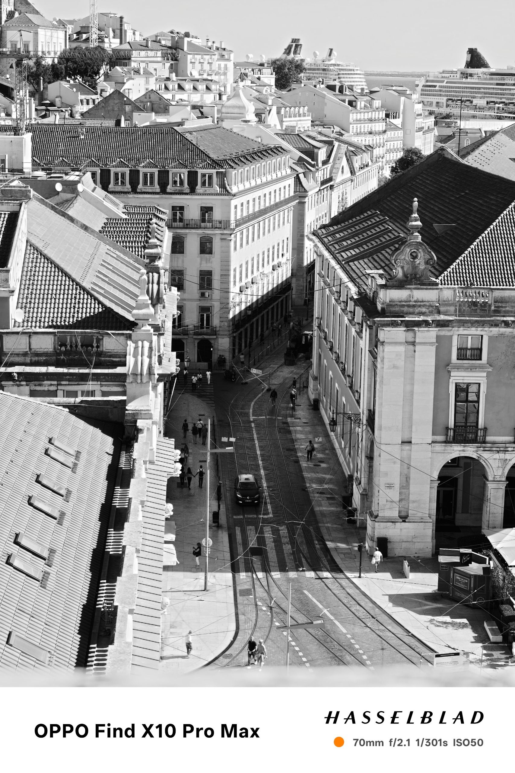 Black-and-white view over Lisbon rooftops and tram-lined streets toward the waterfront.