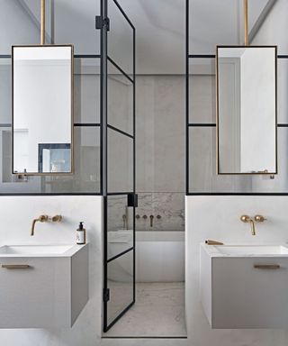 Hotel style bathroom with white marbled flooring, two hand basins, glass door through to white bath, mirrors suspended from the ceiling by copper pipes, copper taps.