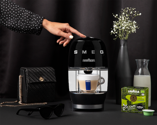Lavazza A Modo Mio Smeg coffee machine in black with woman wearing spotted blouse pressing button, a handbag accessory is in the foreground