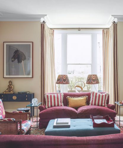 Living room with light yellow painted walls, cherry sofa, lounge chair and blue ottoman, artwork and lampshades in background near bay window