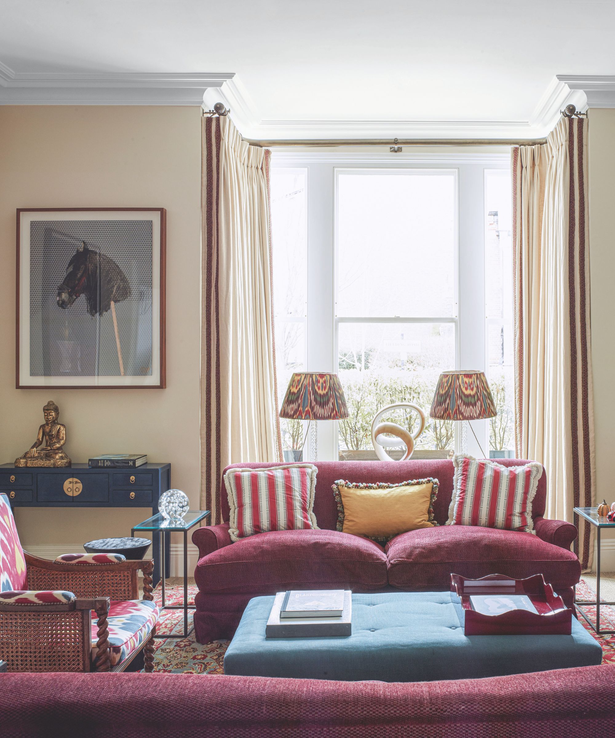 Living room with light yellow painted walls, cherry sofa, lounge chair and blue ottoman, artwork and lampshades in background near bay window