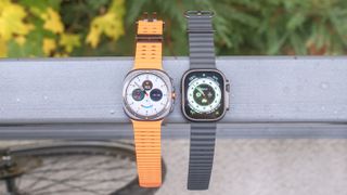 Samsung Galaxy Watch Ultra next to Apple Watch Ultra 2 on a railing with out of focus leaves in the distance