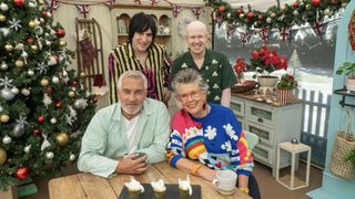 Noel Fielding in a striped shirt, Matt Lucas in a green shirt, Paul Hollywood in a pale green shirt and Prue Leith in a colourful jumper sit in the tent next to a Christmas tree in The Great British Bake Off Christmas special 2022.