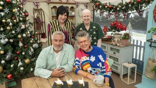 Noel Fielding in a striped shirt, Matt Lucas in a green shirt, Paul Hollywood in a pale green shirt and Prue Leith in a colourful jumper sit in the tent next to a Christmas tree in The Great British Bake Off Christmas special.