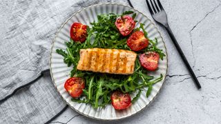 Plate of grilled salmon, high in protein, with green rocket salad and tomatoes on grey plate against grey marble work surface