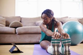A woman sitting on a yoga mat stretches while turning to look at a tablet computer.