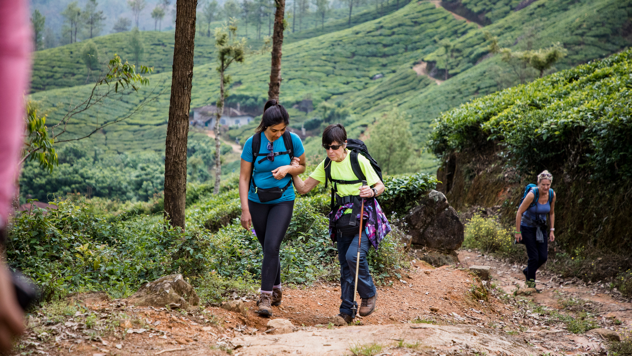 Woman supporting a visually impaired woman on a hike in Munnar, India. They are both smiling and wearing backpacks. The visually impaired woman is using a cane.