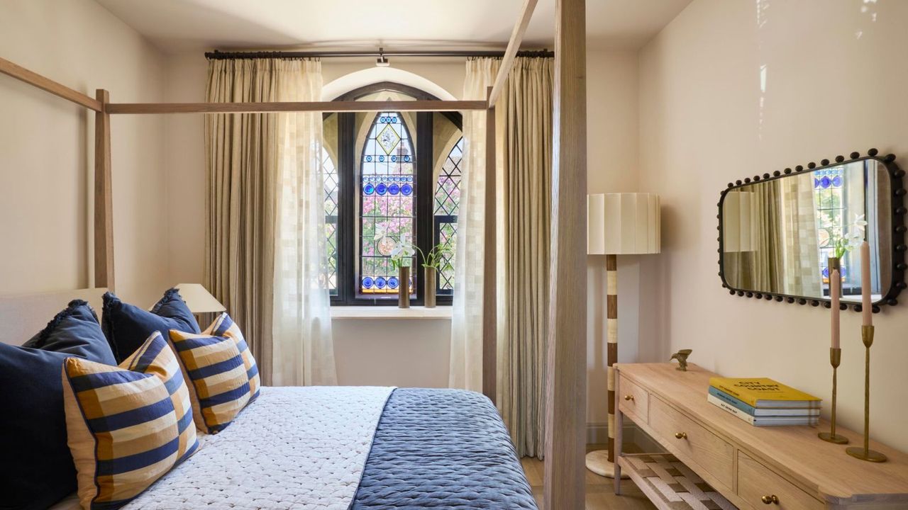 A bedroom inside a chapel conversion with a four-poster bed, blue and yellow cushions, and an original arched stained glass window as the focal point.