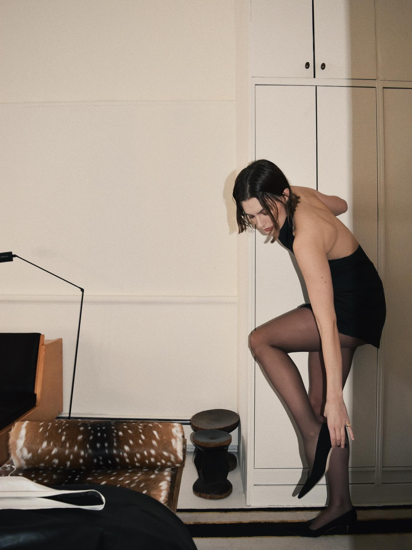 NYC Christie Tyler poses in her chic home wearing a black halter mini dress, black tights, and black kitten heels