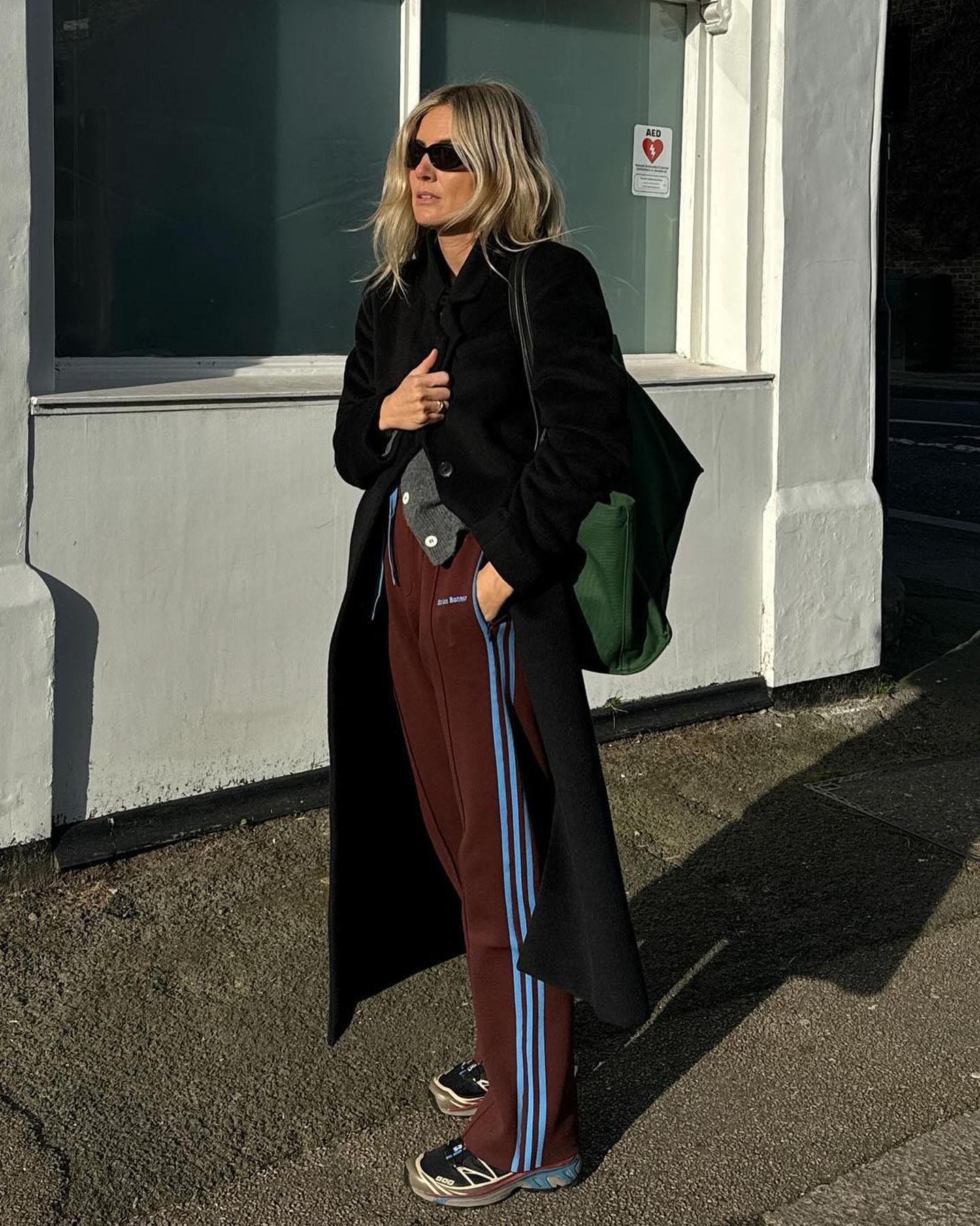 British fashion influencer Lucy Williams poses on the sidewalk in London wearing a long black coat, gray cardigan, Adidas Wales Bonner brown and blue track pants, green The Row tote bag, and Salomon sneakers