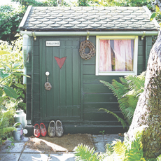 Exterior of a green beach hut summer house with red ginham curtains and shoes outside.