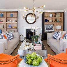 Grey living room with 2 orange chairs and 2 grey couches around a fireplace with brass decorations in the room