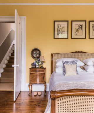 A yellow painted bedroom feature wall idea with antique rattan bed and wooden bedside table and a triptych of paintings above the bed.