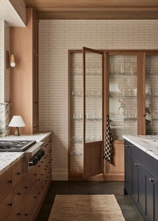 a white tiled wall in a kitchen with glass cabinets fitted into a recess. a dark island can be seen on one side, on the other, a range and small table lamp.