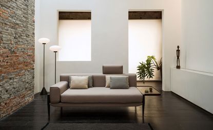A modular upholstered sofa by Michele De Lucchi upholstered in beige fabric and with grey cushions. The sofa is photographed in a room with plants and floor standing lamps, and a brick wall on the left