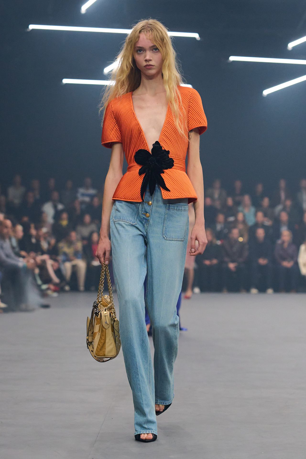 A Valentino model wearing a deep-v-neck orange blouse with a black bow, a pair of light wash baggy jeans, black peep-toe heels, and a tan patchwork handbag with a gold chain strap.