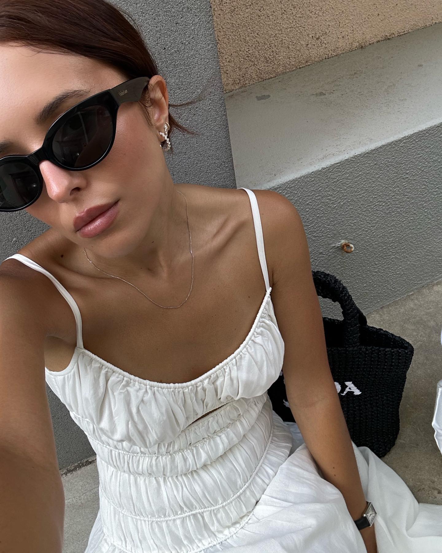 style influencer Debora Rosa poses for selfie wearing black cat eye sunglasses, a black woven Prada tote bag, and white sleeveless smocked dress with thin straps