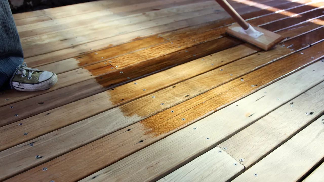 A person wearing denim jeans and casual green and white laced sneakers applying one of the best decking oils to wooden decking with long-handled wooden brush,