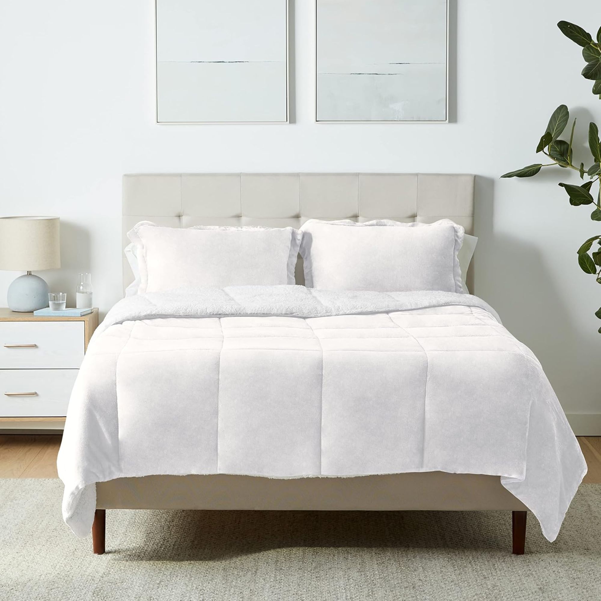 Cream fleecy bedding on a large double bed in a white room. To the left of the bed is a white and wood bedside table with small lamp, and to the right is a large houseplant. Above the bed are two framed white artworks. Beneath the bed is a woven rug, and the flooring is wood.