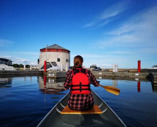 a person's back is seen. they are holding an ore sitting at the front of a canoe. water surrounds. nearby a constructed shoreline.