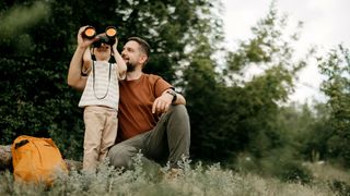 A picture of a man helping a little boy look through his binoculars whilst on a trip outdoors