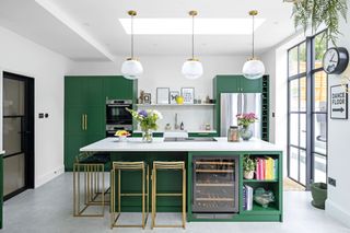Green modern kitchen with island; white quartz worktop, brass bar stools, brass and glass pendant lights and polished concrete floor
