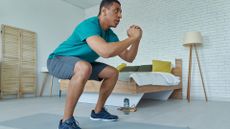 man in a turquoise tshirt and grey shorts squatting indoors with a bed and lamp behind him facing sideways to the camera