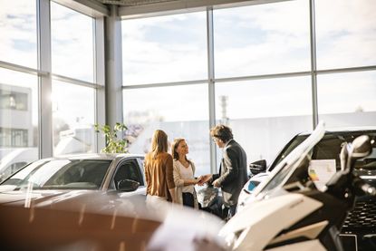 Salesman is giving keys to a female customer, both smiling at each other. Another woman is standing nest to her, indoors at a car dealership.