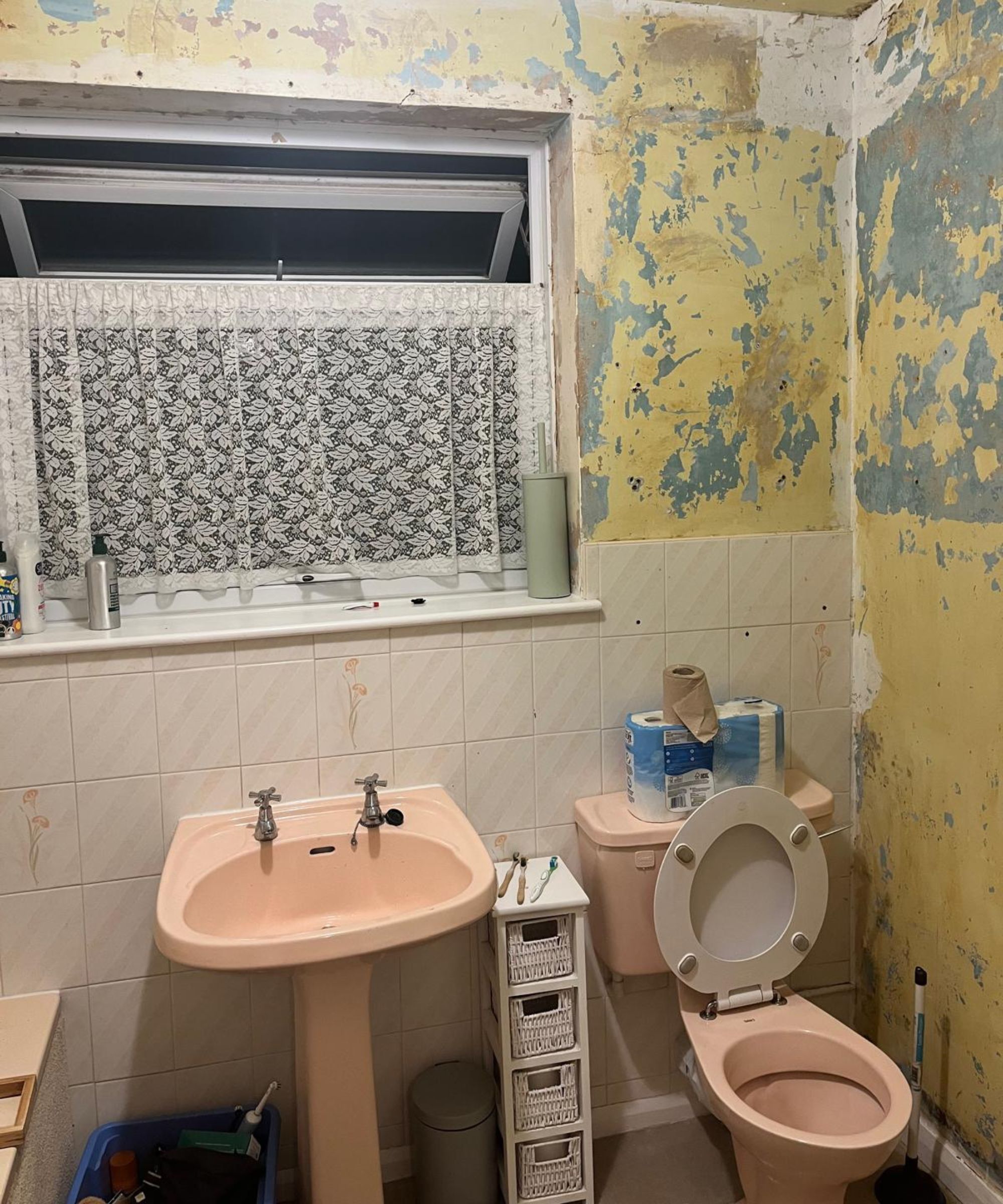 A dated bathroom with a pink suite, heavily peeling yellow walls and old floral tiles.