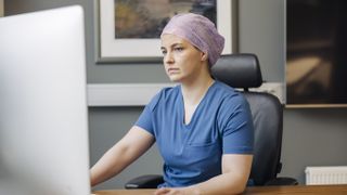 white woman wearing blue medical scrubs and a surgeon's head covering sits at a desktop computer as if reviewing patient data 