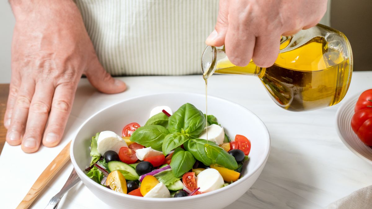 Person pouring olive oil from a jug onto a bowl of salad which includes basil, tomatoes, black olives and mozzarella