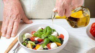 Person pouring olive oil from a jug onto a bowl of salad which includes basil, tomatoes, black olives and mozzarella