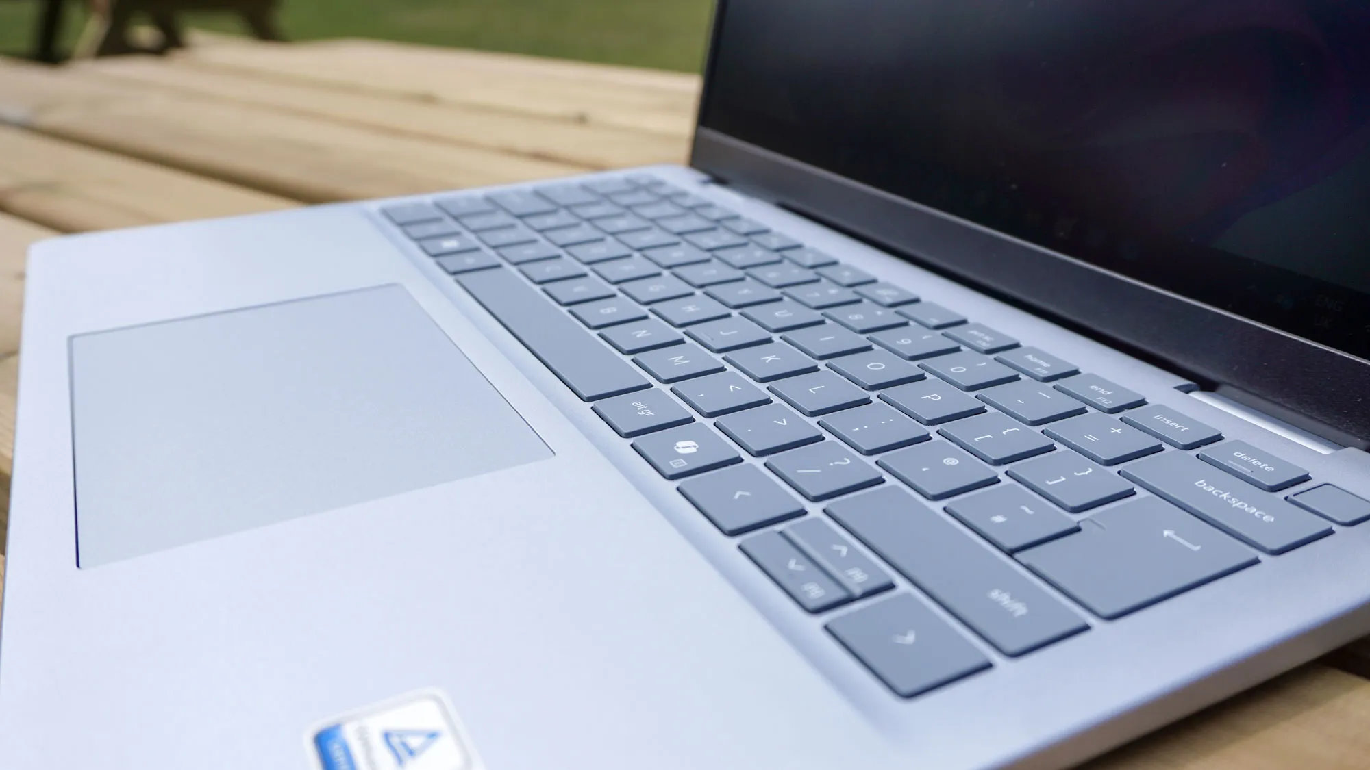 A sleek silver Dell 14 Plus laptop on a wooden table, viewed from a side angle. The focus is on the keyboard and touchpad, showcasing a clean, modern design.