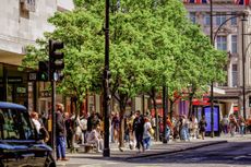  A view along Oxford Street, looking towards Oxford Circus, with the University of the Arts London: London College of Fashion building visible on the left. The street is lined with green trees, and various shops line the pavement, creating a vibrant scene with pedestrians and shoppers nearby.